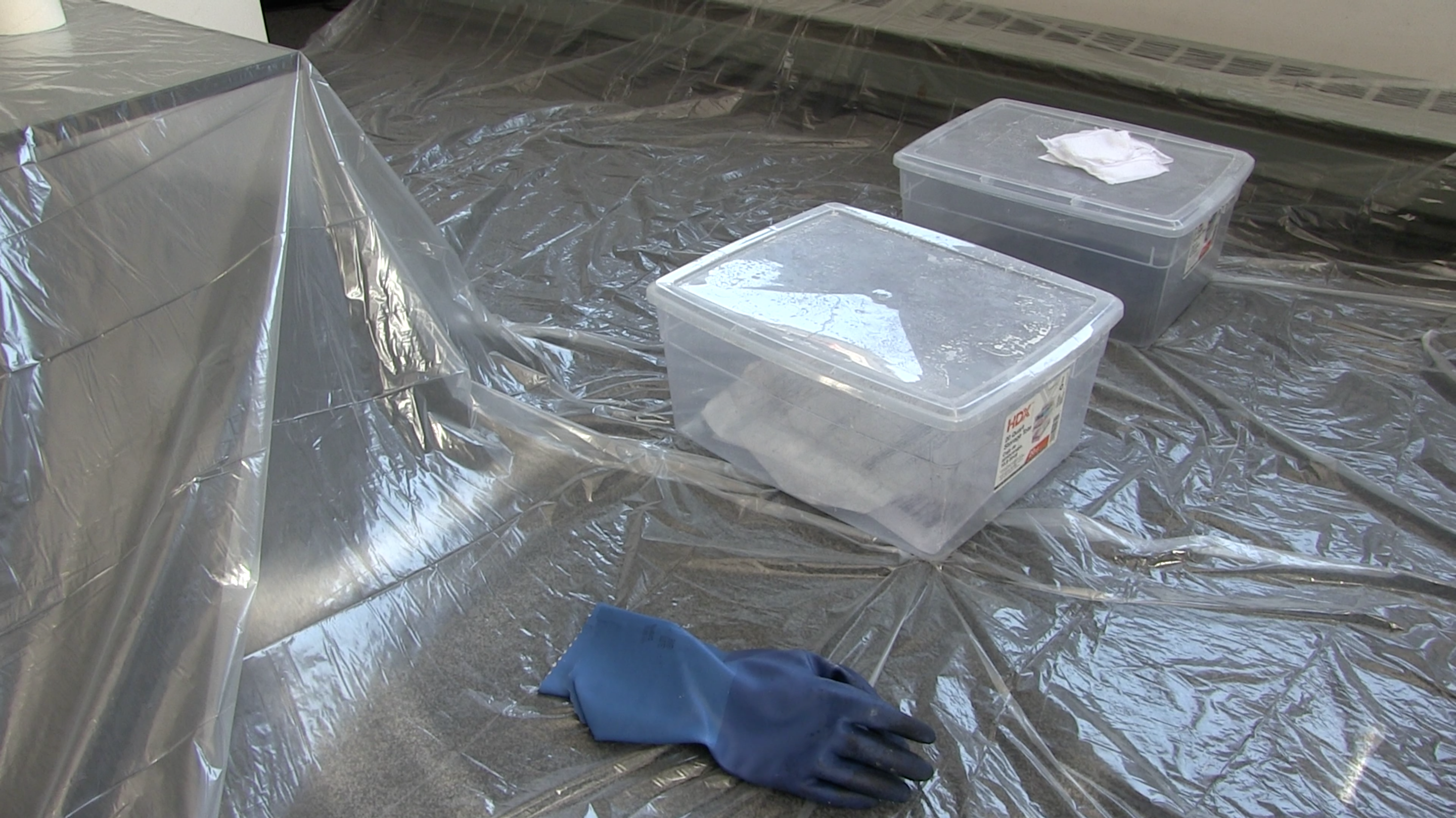 Floor covered in a clear plastic, two closed bins with liquid and floating documents inside, and a pair of blue gloves.