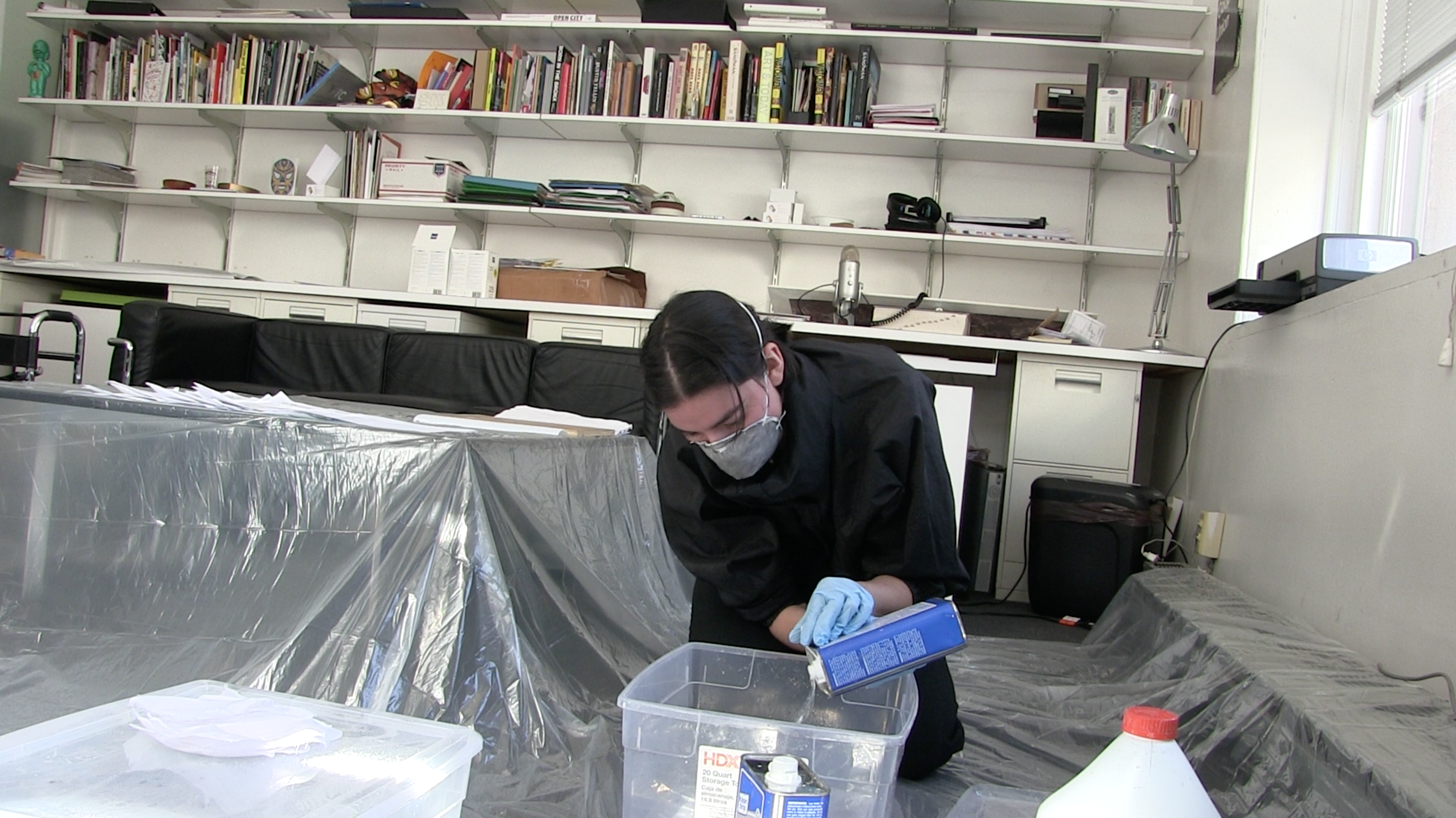 Performer on the floor pouring a chemical into one of the bins. They wear a gas mask and blue gloves.