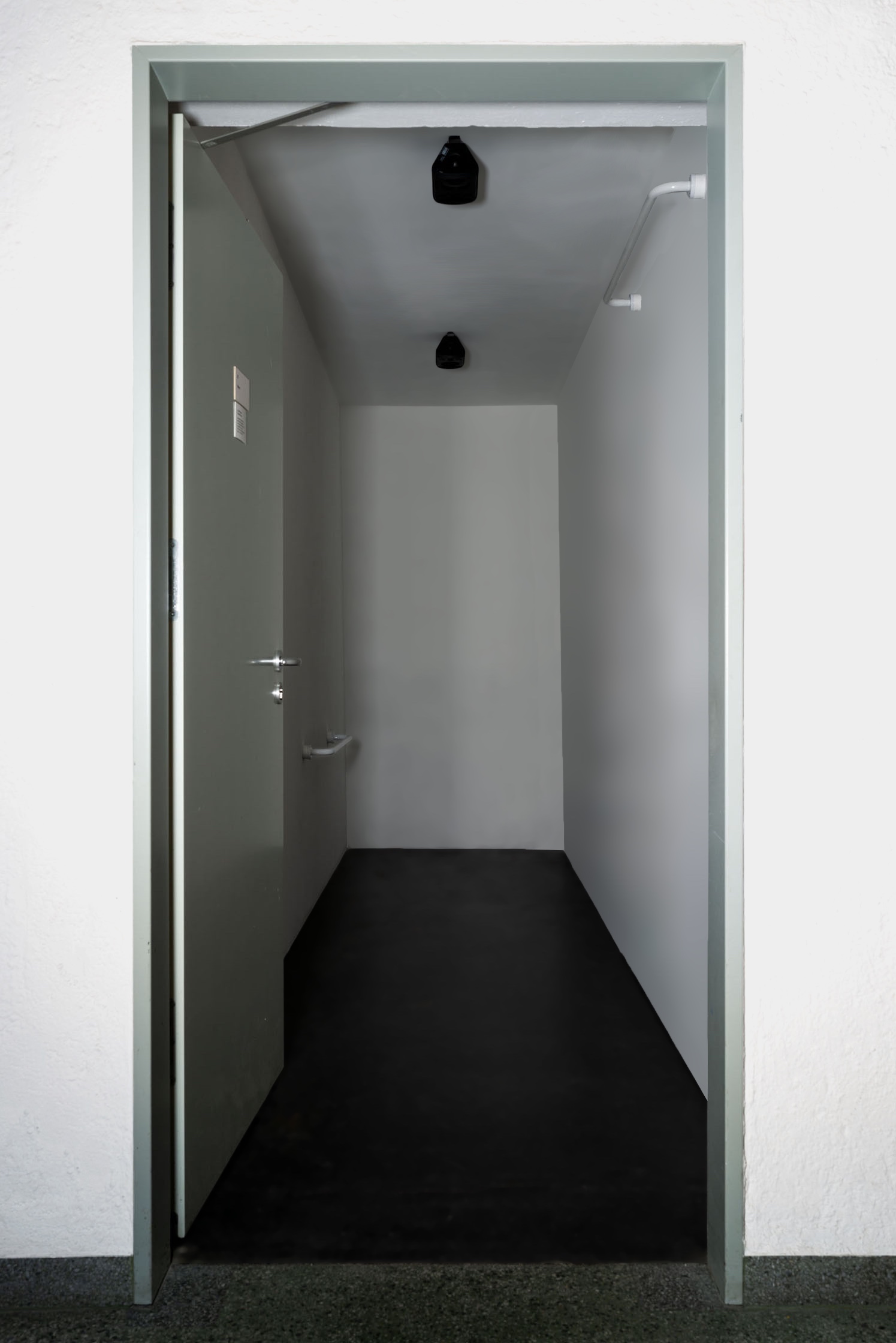 Outer view of built in room, door propped open, revealing a long narrow white box room. Two speakers mounted equidistant on the ceiling of the room. Flagged by two accessibility handles on opposite cornes of the left and right wall. 