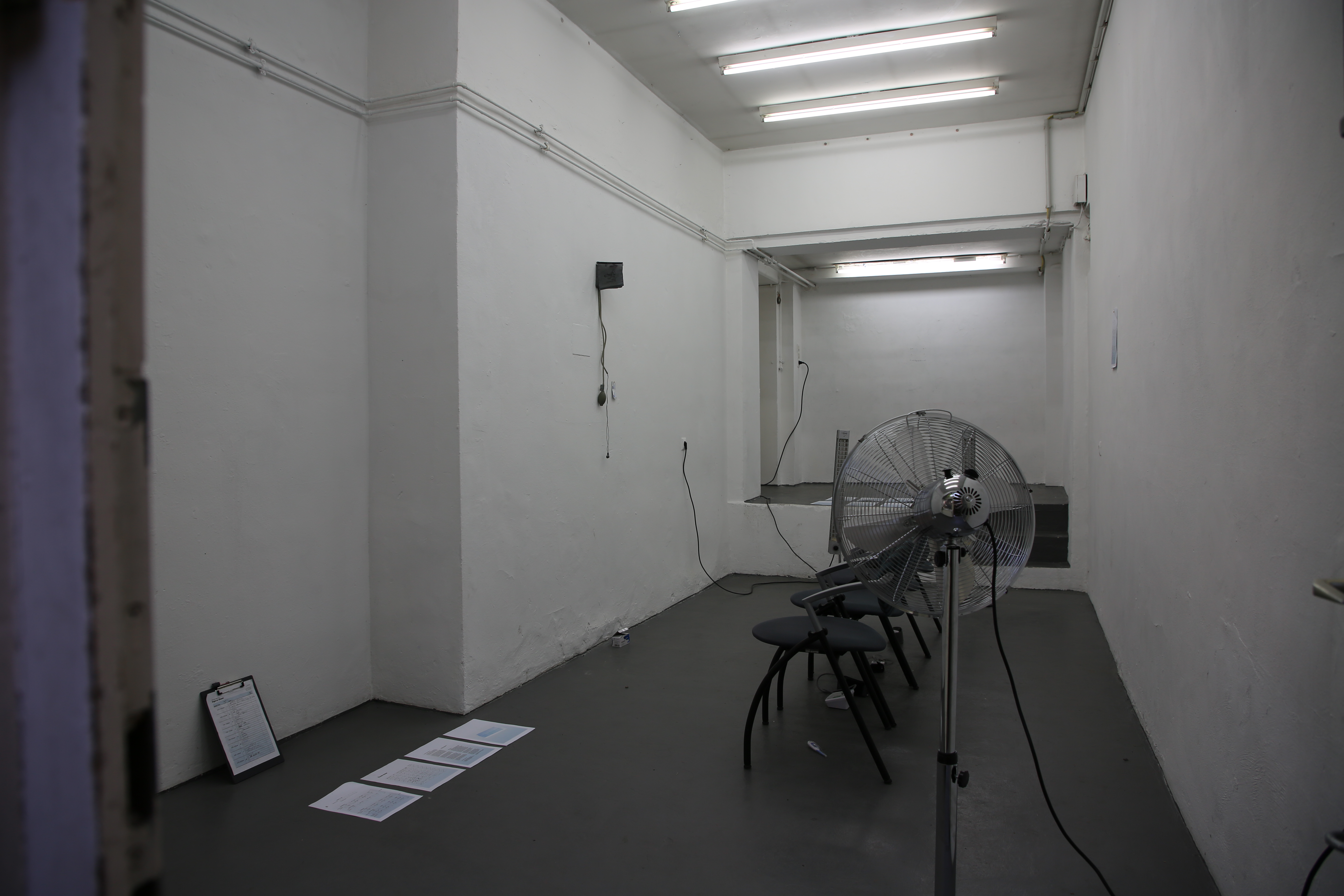 Installation view, One fan faces away from entrance door into the room. 4 documents sit on left side of the floor. Clipboard against the wall. Down the center of the room are 4 waiting room chairs facing the left wall. A blood pressure arm wrap hangs on this wall with two headphones emerging from its connection tubes. in the back are two heaters facing the entrance wall.